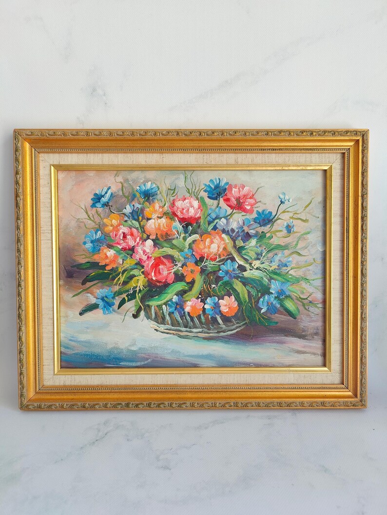 May include: Oil painting of a floral arrangement in a basket, framed in a gold-colored ornate frame. The flowers are a mix of pink, orange, and blue, with green leaves. The background is a soft, light color.