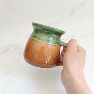 May include: A ceramic mug with a glossy green rim and a textured, speckled brown body. The mug features a decorative beaded band detail below the rim and a small stamped logo on the side that reads "DENBY" and "1809". The mug has a wide base.