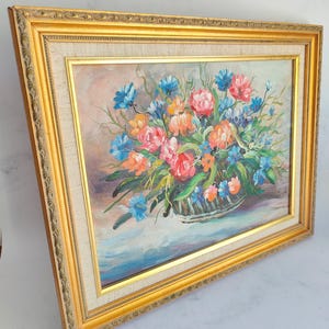 May include: A framed floral oil painting featuring a basket overflowing with colorful flowers. The artwork showcases pink, orange, and blue blooms, set against a soft background. The painting is encased in a gold ornate frame.