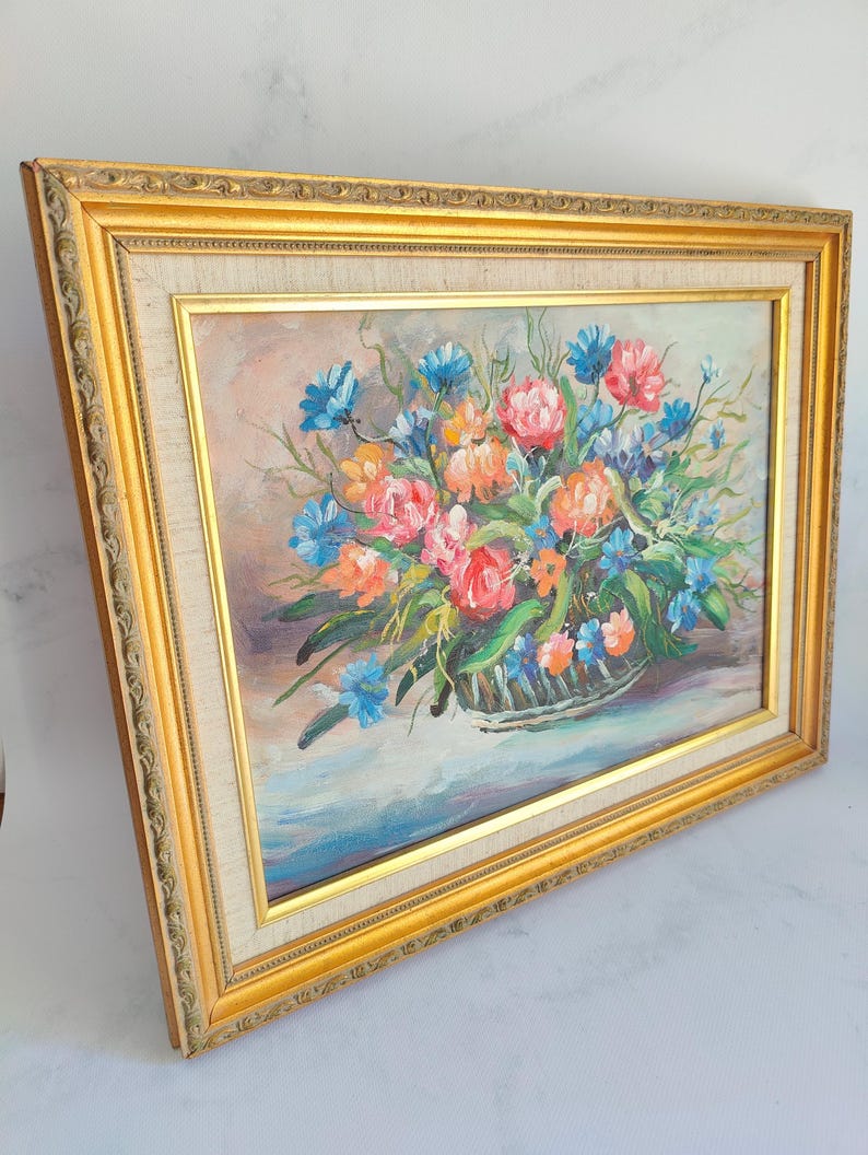 May include: A framed floral oil painting featuring a basket overflowing with colorful flowers. The artwork showcases pink, orange, and blue blooms, set against a soft background. The painting is encased in a gold ornate frame.