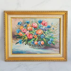 May include: Oil painting of a floral arrangement in a basket, framed in a gold ornate frame. The flowers are a mix of pink, orange, and blue, with green foliage. The background is a soft, neutral color.