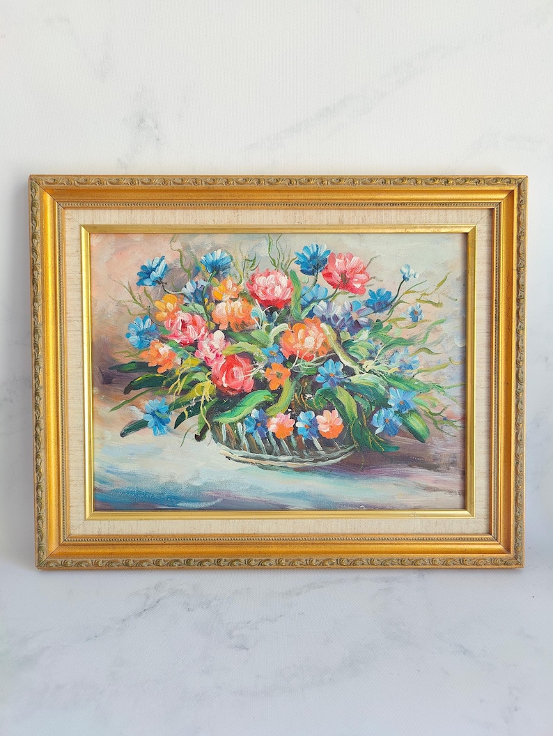 May include: Oil painting of a floral arrangement in a basket, framed in a gold ornate frame. The flowers are a mix of pink, orange, and blue, with green foliage. The background is a soft, neutral color.
