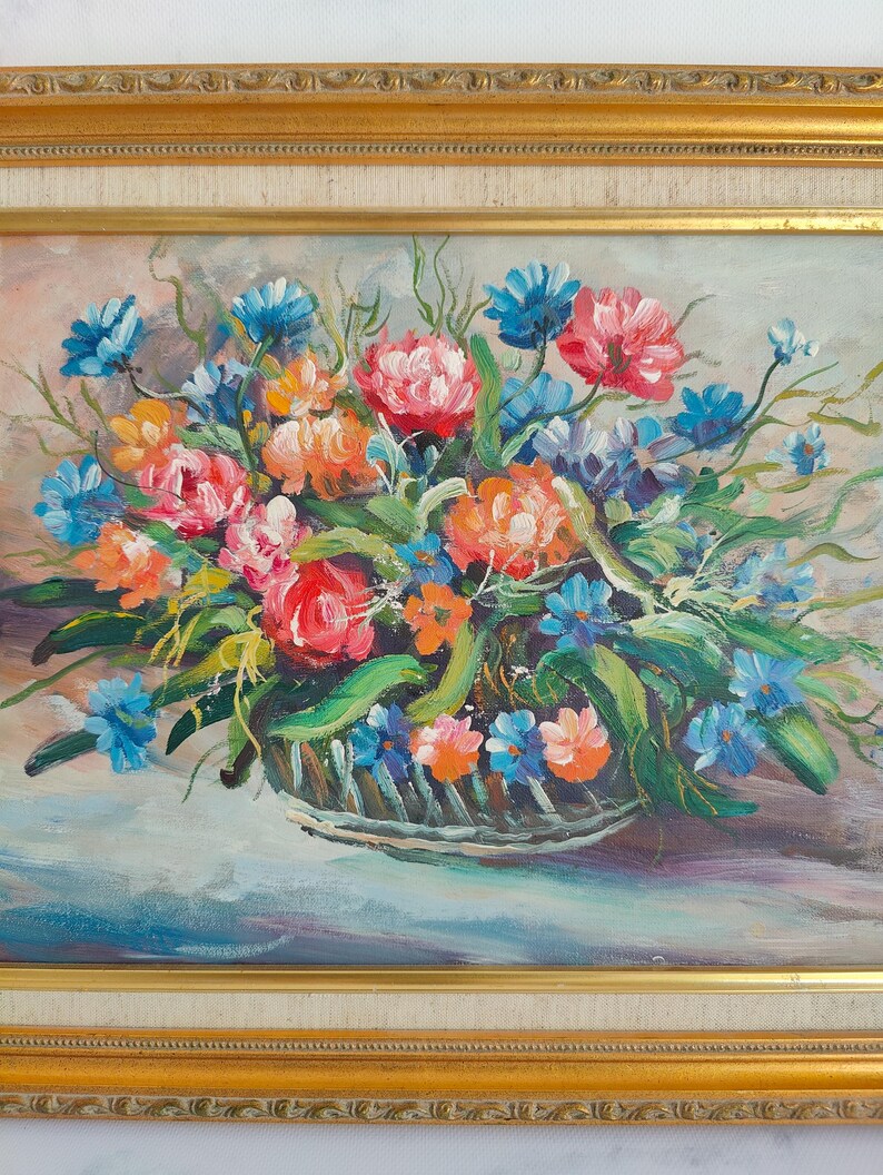 May include: Oil painting of a floral arrangement in a basket, framed in gold. The flowers are a mix of red, orange, and blue, with green leaves. The background is a soft, muted color.