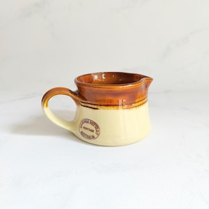 May include: A small, ceramic creamer with a handle and a spout. The creamer is two-toned, with a light beige base and a brown upper section. The handle and the interior are also brown. The text "Ringo Pottery Heritage Australia" is printed on the side.