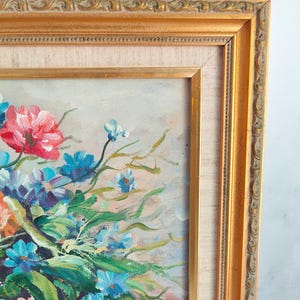May include: Oil painting of a floral arrangement in a gold ornate frame. The bouquet features red, blue, and orange flowers with green leaves. The frame has a detailed, textured design.