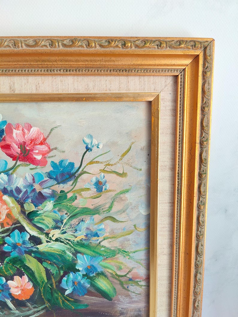 May include: Oil painting of a floral arrangement in a gold ornate frame. The bouquet features red, blue, and orange flowers with green leaves. The frame has a detailed, textured design.