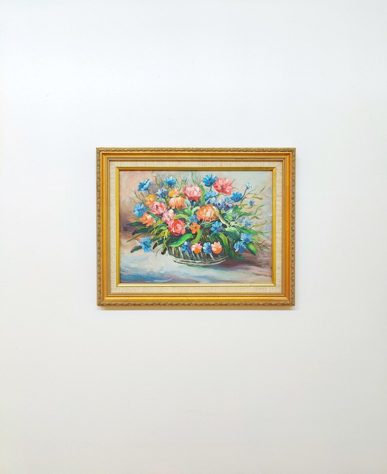 May include: A framed floral oil painting featuring a vibrant bouquet of pink, orange, and blue flowers. The flowers are arranged in a decorative basket, set against a soft, light background. The frame is gold-colored with ornate detailing.