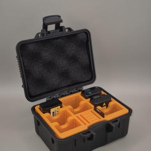 Könnte beinhalten: Schwarzer und orangefarbener Hartschalenkoffer mit offenem Deckel. Der Innenraum hat eine schwarze Schaumstoffauskleidung im Deckel und orangefarbene Fächer im Boden. Mehrere kleine elektronische Geräte sind im Inneren verstaut.