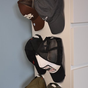 Könnte beinhalten: Fünf Baseballkappen hängen an einer weißen Wand. Die Kappen sind in verschiedenen Farben und Stilen, darunter Braun, Schwarz und Grün. Einige der Kappen haben Netzpanels und Logos.