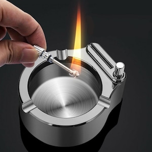 May include: A silver ashtray with a built-in lighter. The lighter is being used to light a match. The ashtray has a round, deep bowl and a lid with a built-in lighter.