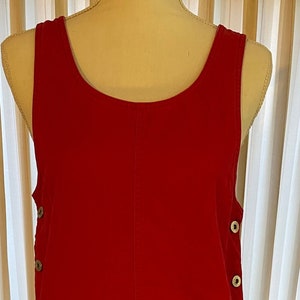 May include: A red sleeveless corduroy overall dress with a scoop neck and two pockets. The dress has silver buttons on the sides.
