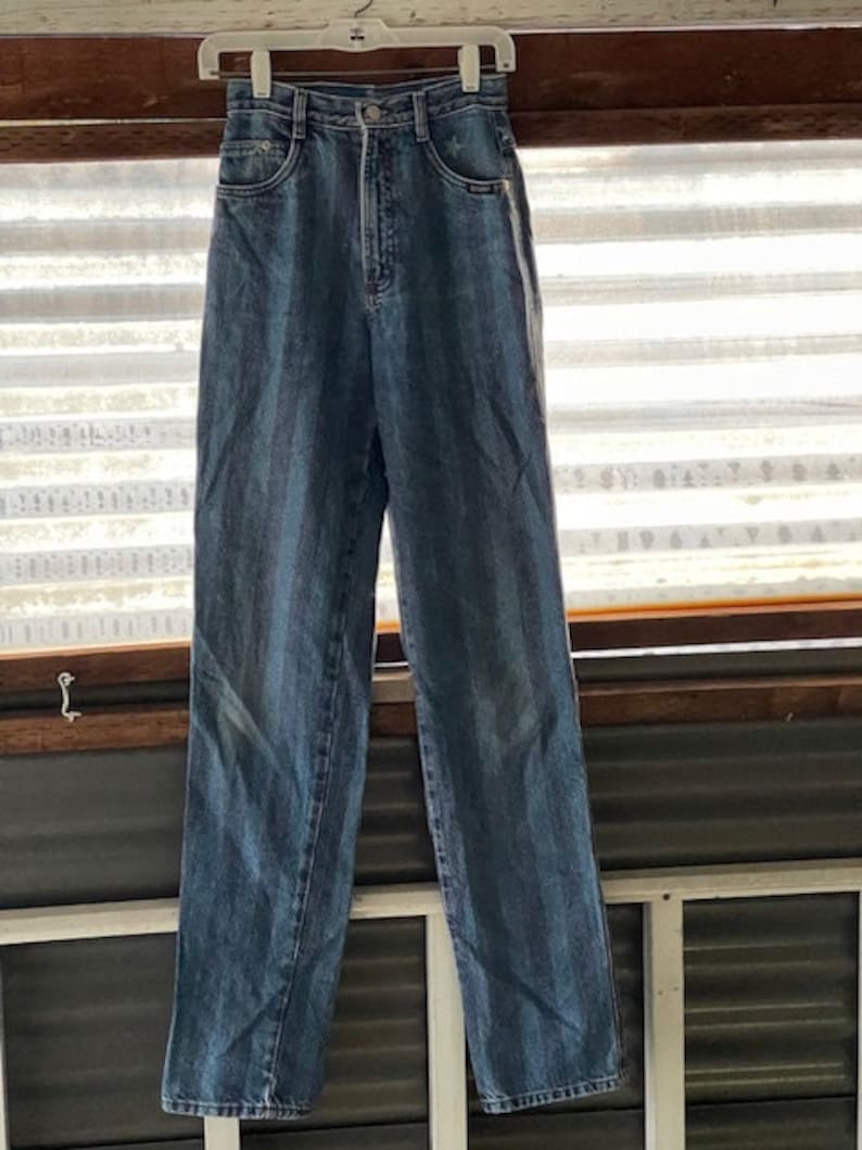 May include: A pair of blue denim jeans with a white stripe down the side seams. The jeans are hanging on a white hanger.