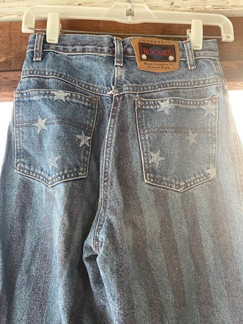 May include: Blue denim jeans with a white star pattern and a brown leather patch that says "Authentic Rockies World Champion Fit from the Denim City".