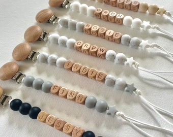 Pode incluir: Um conjunto de sete mordedores de contas de madeira com diferentes nomes escritos neles. Os mordedores são feitos com contas de madeira natural e contas de silicone em cinza, branco e preto. Cada mordedor tem um clipe de madeira anexado.