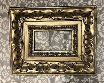 May include: A gold-colored ornate picture frame with a decorative, raised design. The frame has a rectangular opening for a picture or artwork.