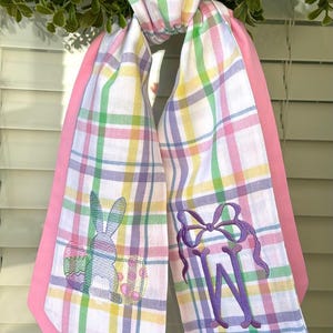 May include: A plaid scarf with pink edging, featuring embroidered Easter-themed designs. One side has a bunny and an egg, the other a purple monogram. The scarf is white with a pastel plaid pattern of pink, yellow, green, and purple.