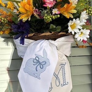 May include: A vibrant floral display featuring daisies, yellow and pink flowers. Two white fabric banners are attached, one with a blue embroidered bunny and the other with a blue embroidered letter "M".