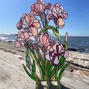 May include: A stained glass panel featuring a cluster of pale pink iris flowers with deep red accents and vibrant green stems and leaves. The piece is held upright by a light-coloured wooden stand, showcasing intricate glass soldering and textures.