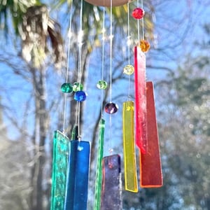 May include: A wind chime featuring a circular wooden ring from which hang multiple translucent, rectangular glass pieces in various colors including red, yellow, green, blue, and purple, accented with small, colorful glass beads on thin strings.