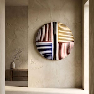 Op de afbeelding: Een groot, rond wandkunstwerk met een geometrisch ontwerp. Het kunstwerk is verdeeld in vier secties, elk met verticale strepen in rood, geel, blauw en zwart, gescheiden door witte strepen. Het stuk is bevestigd aan een getextureerde, beige muur.
