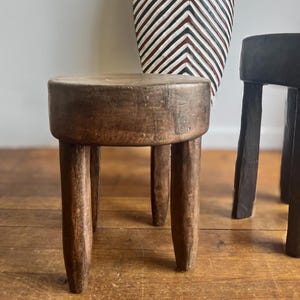 Puede incluir: Un pequeño taburete redondo de madera con tres patas. El taburete está hecho de madera de color marrón oscuro y tiene un aspecto rústico y envejecido. El taburete es un objeto decorativo.