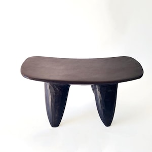 Puede incluir: Un taburete de madera marrón oscuro con tres patas. El taburete tiene una parte superior curva y un acabado liso y pulido.