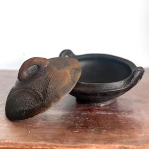 Peut inclure: Un petit pot rond en terre cuite noire avec un couvercle. Le couvercle a une poignée et est décoré d'un motif. Le pot est posé sur une surface en bois.