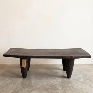 Op de afbeelding: Een donkerbruine houten salontafel met een gebogen rechthoekig blad en vier dikke, taps toelopende poten. Aan een poot hangt een label en rechtsonder staat de tekst: BKwo3 - Longueur 1m08cm x H 30cm x l 35cm.