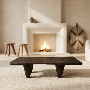 May include: A dark brown, rectangular coffee table with four thick, tapered legs. The table has a textured surface and sits on a light-colored carpet. The background includes a fireplace with a burning fire, two small wooden stools, and decorative items.