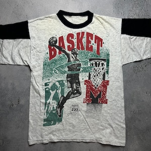 Peut inclure: Un t-shirt gris et noir avec un motif de joueur de basket-ball. Le motif représente un joueur en plein saut, en train de dunker un ballon de basket. Le texte "BASKET" est imprimé en rouge au-dessus du joueur, et le texte "M" est imprimé en rouge en dessous du joueur. Le texte "233 STREET WEAR" est imprimé en rouge et blanc en dessous du joueur.