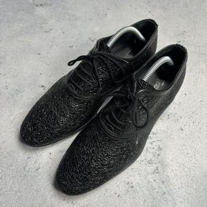 Może przedstawiać: Para czarnych sk&oacute;rzanych but&oacute;w wizytowych z teksturowanymi detalami. Buty mają klasyczny design sznurowany i są prezentowane na szarej powierzchni. Wewnątrz but&oacute;w umieszczono prawidła, aby zachować ich kształt.