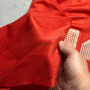 Puede incluir: Primer plano de una camiseta roja con un patr&oacute;n texturizado. La camiseta tiene un dise&ntilde;o blanco y beige en la parte inferior derecha, con un emblema de gallo. La manga est&aacute; doblada, revelando el interior de la tela.