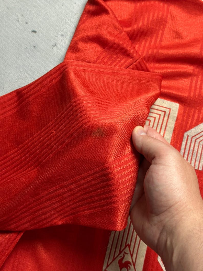 Puede incluir: Primer plano de una camiseta roja con un patr&oacute;n texturizado. La camiseta tiene un dise&ntilde;o blanco y beige en la parte inferior derecha, con un emblema de gallo. La manga est&aacute; doblada, revelando el interior de la tela.