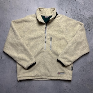 Könnte beinhalten: Ein beiger Fleece-Pullover mit einem halben Reißverschluss und schwarzen Einfassungen. Der Reißverschluss hat einen silbernen Zipper und das Fleece hat eine weiche, flauschige Textur. Der Markenname Berghaus ist auf der rechten unteren Seite des Kleidungsstücks sichtbar.