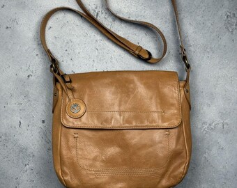 May include: A tan leather crossbody bag with a flap closure and adjustable strap. The bag features a small, circular emblem on the front and a stitched design. The bag's strap is approximately 40 inches long.