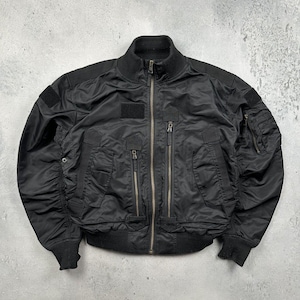 May include: Black bomber jacket with a ribbed collar, cuffs, and hem. The jacket features multiple pockets with zipper closures and a patch on the chest. The jacket has a full-length zipper and a classic bomber silhouette.