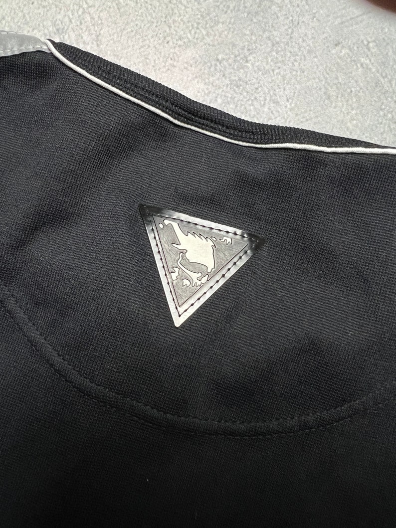 May include: Black t-shirt with a white trim around the neckline. A silver triangle-shaped patch with a design is sewn onto the back of the shirt.