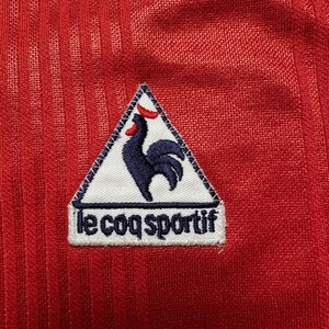 Puede incluir: Primer plano de una camiseta deportiva roja con un patr&oacute;n texturizado y acanalado. Un parche blanco con las palabras "le coq sportif" est&aacute; cosido en la camiseta. El parche presenta un gallo azul marino con cresta y pico rojos.