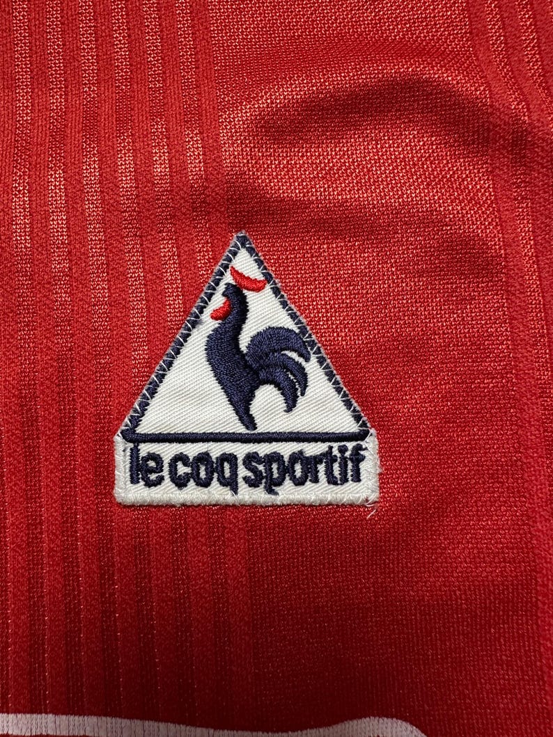 Puede incluir: Primer plano de una camiseta deportiva roja con un patr&oacute;n texturizado y acanalado. Un parche blanco con las palabras "le coq sportif" est&aacute; cosido en la camiseta. El parche presenta un gallo azul marino con cresta y pico rojos.