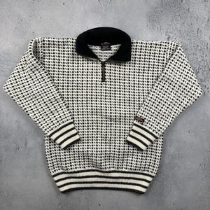May include: A black and white patterned sweater with a black collar and a quarter-zip closure. The cuffs and hem feature black and white striped detailing. The sweater has a textured appearance and a classic design.