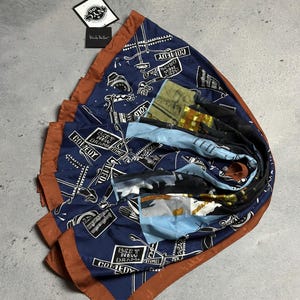 Puede incluir: Una bufanda de seda azul con un borde marrón. La bufanda presenta un estampado de una marquesina de cine con las palabras "Comedy", "Drama", "Best New" y "Times" impresas en ella. La bufanda también presenta un estampado de un carrete de película y una cámara de cine.