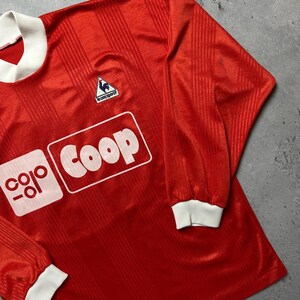 Puede incluir: Una camiseta de f&uacute;tbol roja de manga larga con pu&ntilde;os y cuello blancos. La camiseta presenta el texto "coop" en un recuadro blanco y un peque&ntilde;o logotipo de gallo. La tela tiene rayas verticales.