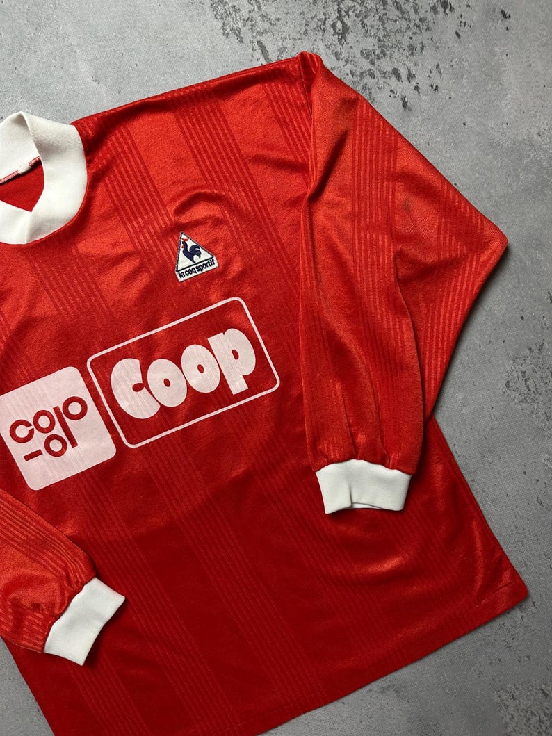 Puede incluir: Una camiseta de f&uacute;tbol roja de manga larga con pu&ntilde;os y cuello blancos. La camiseta presenta el texto "coop" en un recuadro blanco y un peque&ntilde;o logotipo de gallo. La tela tiene rayas verticales.