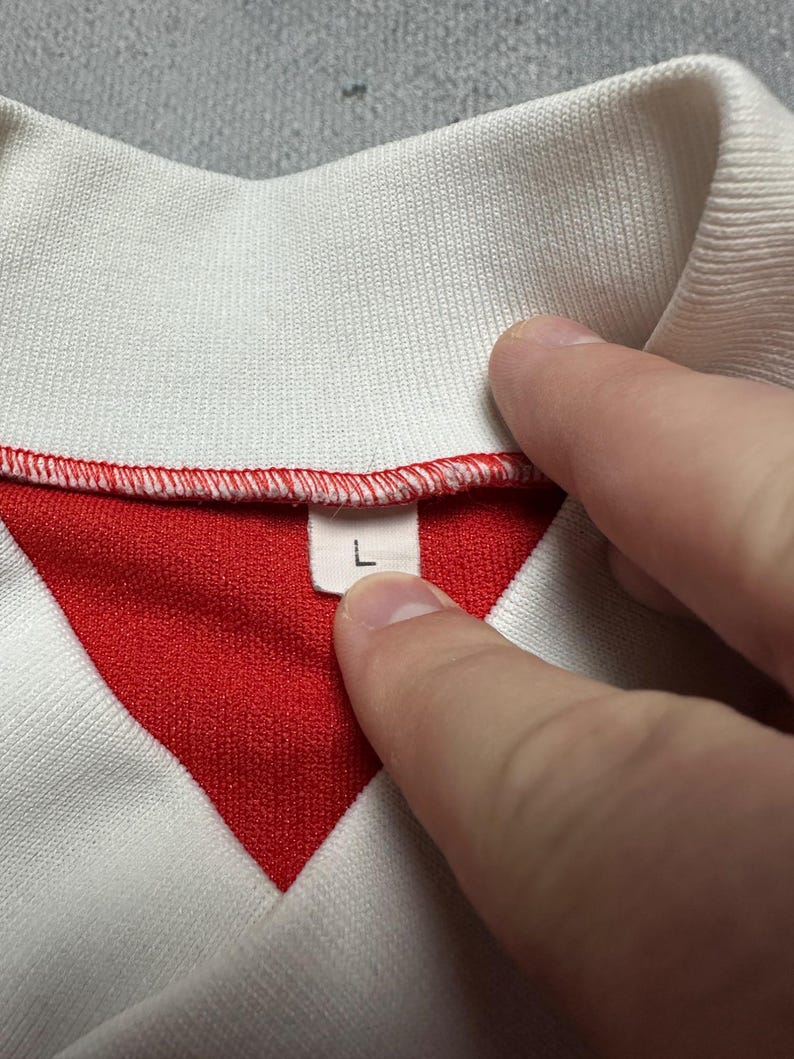 Puede incluir: Primer plano de un cuello de camisa blanco y rojo. El cuello es blanco, con el interior rojo. La etiqueta de talla muestra la letra "L". La camisa est&aacute; hecha de una tela suave y texturizada.