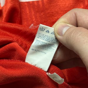 Puede incluir: Primer plano de una tela roja con una etiqueta blanca. La etiqueta tiene instrucciones de lavado y el texto "100% POLYESTER". La tela es texturizada y parece ser parte de una prenda.