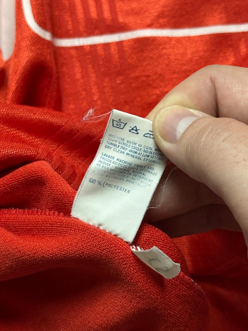 Puede incluir: Primer plano de una tela roja con una etiqueta blanca. La etiqueta tiene instrucciones de lavado y el texto "100% POLYESTER". La tela es texturizada y parece ser parte de una prenda.