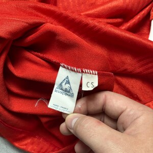 Puede incluir: Primer plano de una camiseta deportiva roja con rayas blancas y una etiqueta Le Coq Sportif. La etiqueta dice "PARIS - FRANCE" y presenta un logotipo de gallo. La camiseta est&aacute; hecha de una tela texturizada.