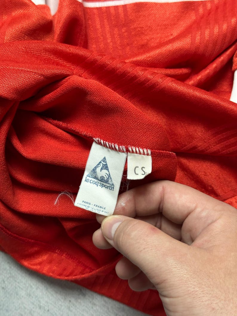 Puede incluir: Primer plano de una camiseta deportiva roja con rayas blancas y una etiqueta Le Coq Sportif. La etiqueta dice "PARIS - FRANCE" y presenta un logotipo de gallo. La camiseta est&aacute; hecha de una tela texturizada.
