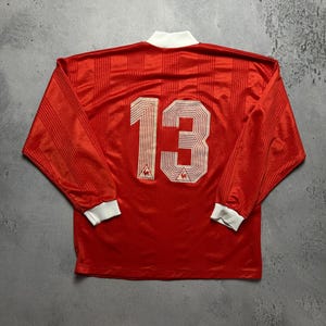 Puede incluir: Camiseta deportiva roja de manga larga con el n&uacute;mero 13 en la espalda. La camiseta tiene cuello y pu&ntilde;os blancos. El logotipo de Le Coq Sportif es visible debajo del n&uacute;mero.
