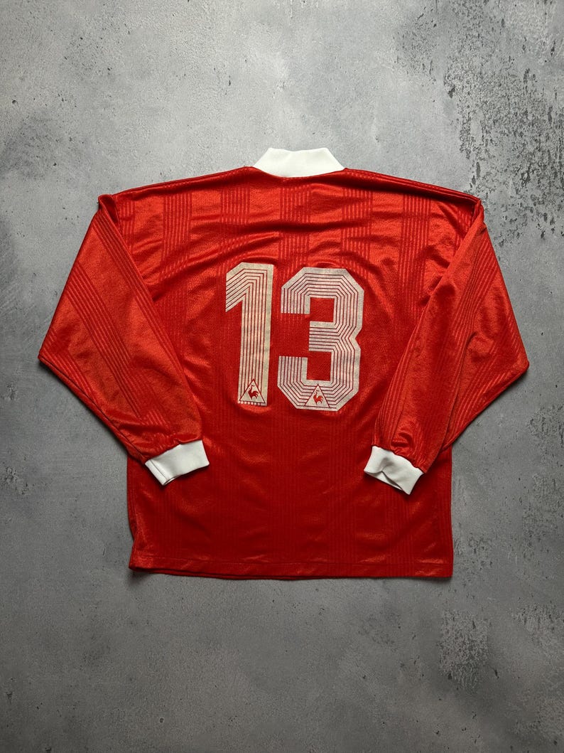 Puede incluir: Camiseta deportiva roja de manga larga con el n&uacute;mero 13 en la espalda. La camiseta tiene cuello y pu&ntilde;os blancos. El logotipo de Le Coq Sportif es visible debajo del n&uacute;mero.